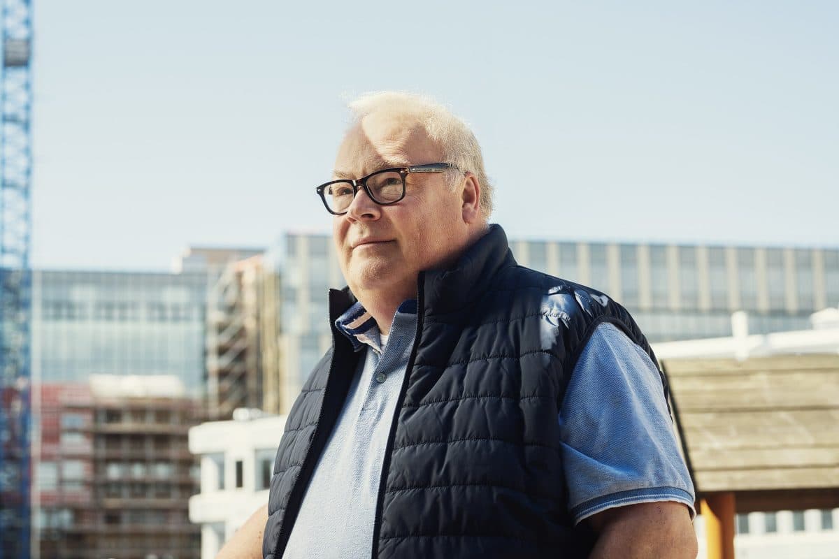 En eldre mann med briller og marineblå vattert vest står utendørs foran moderne kontorbygninger på en solrik dag.