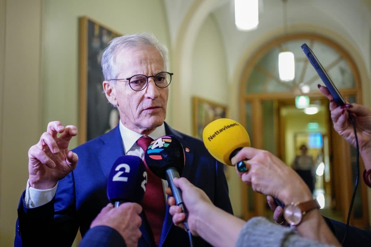 En mann i dress og briller snakker med flere reportere som holder mikrofoner merket med ulike nyhetslogoer i en korridor.