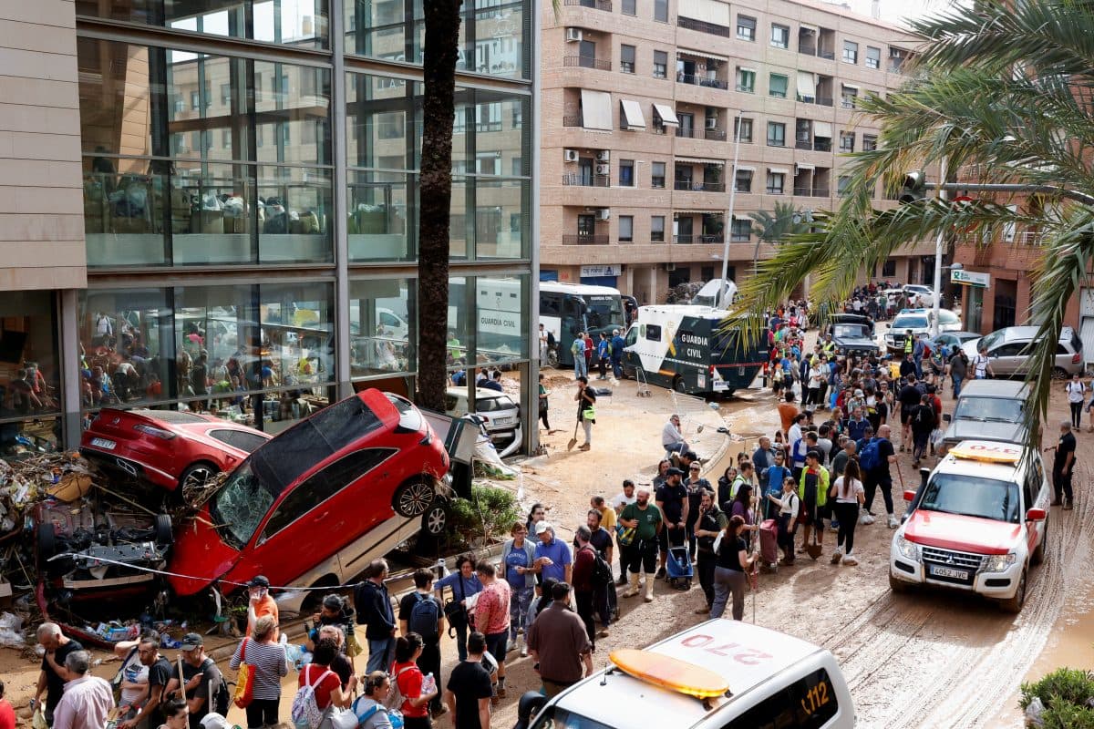 En stor folkemengde samler seg i nærheten av ødelagte biler og utrykningskjøretøy utenfor bygninger, med vrakrester på bakken, etter en ødeleggende hendelse i et byområde.