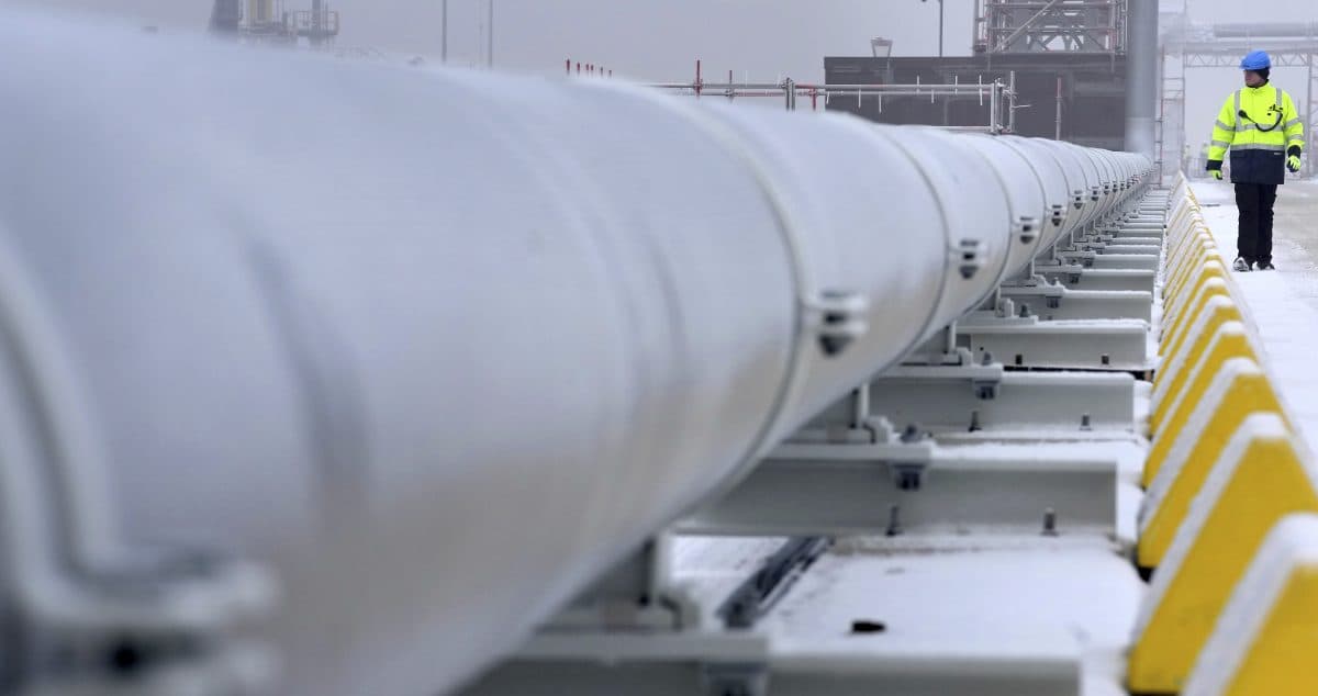 En arbeider iført varseljakke og hjelm går langs en stor industrirørledning en snørik dag.