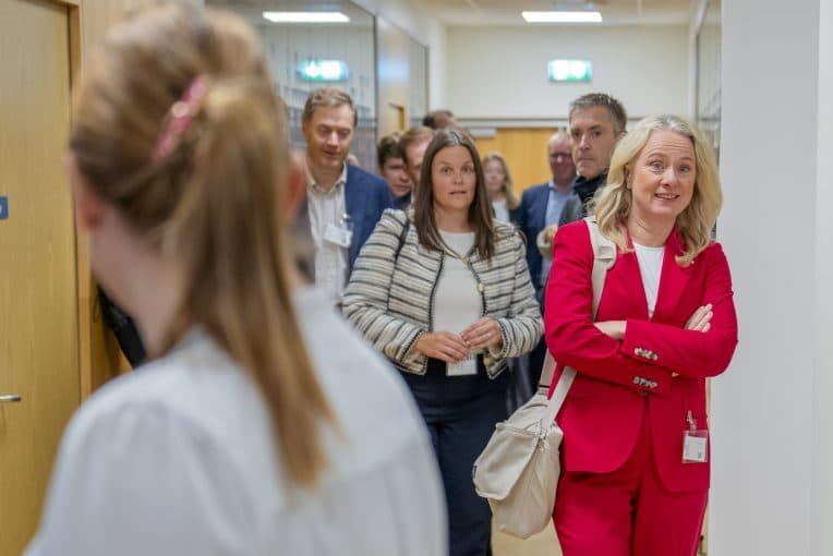 En gruppe mennesker går ned en gang; en kvinne i rød dress med skilt og hvite sko står i forgrunnen og ser fremover med armene i kors.