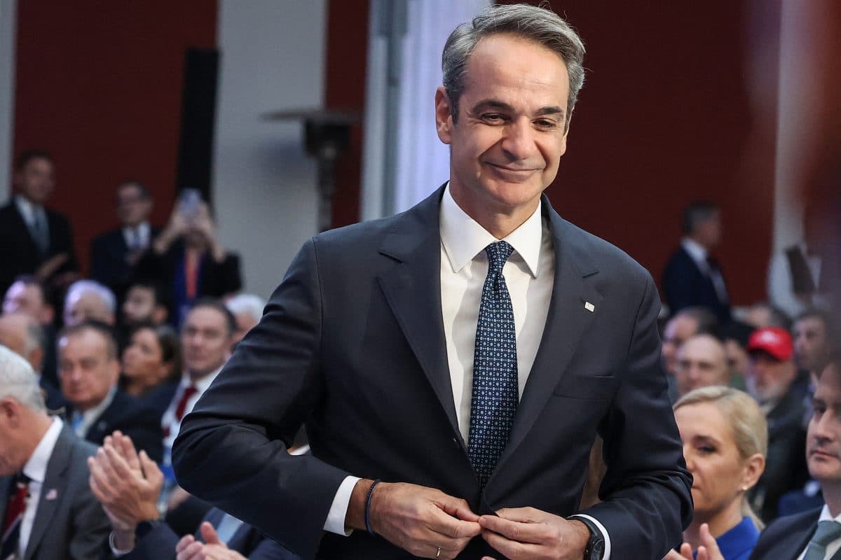 En mann i dress knepper jakka mens han står blant sittende mennesker på et formelt innendørsarrangement.