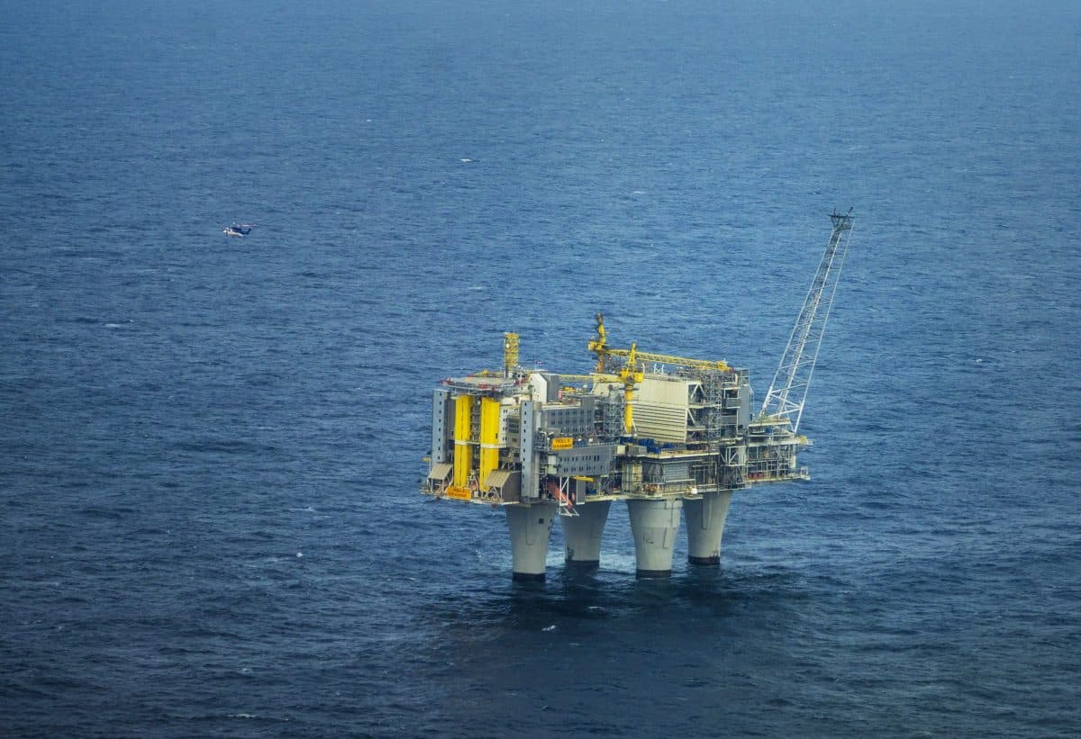 Oljeplattformen står i havet med kraner og utstyr synlig; et helikopter nærmer seg i det fjerne fra venstre side.