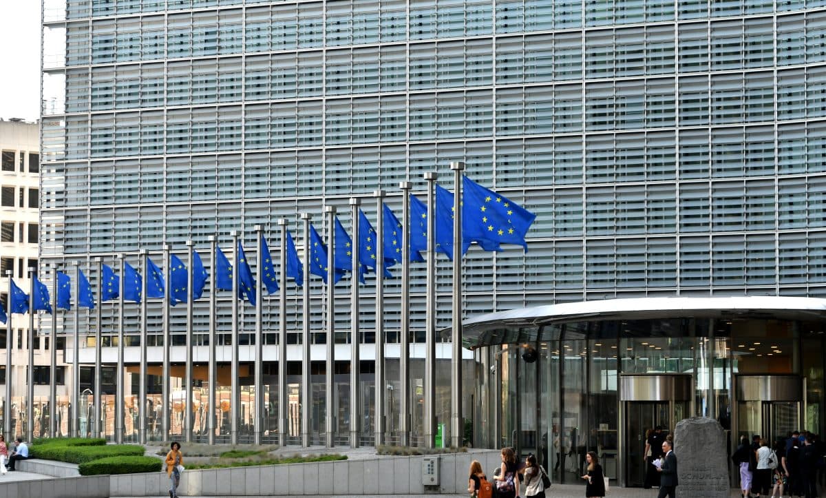 En rekke EU-flagg står utstilt foran en moderne glassbygning mens folk går i nærheten.