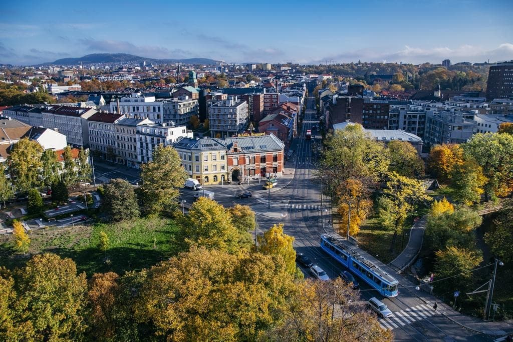Flyfoto av et bykryss med en blå trikk, omkringliggende høsttrær og bygninger under en klar himmel.