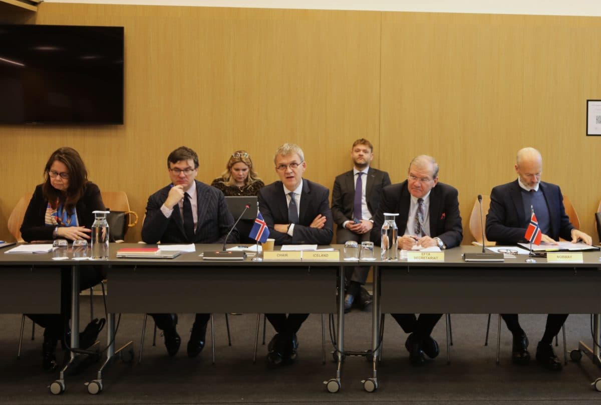 Seks personer i forretningsantrekk sitter ved et konferansebord med navneskilt, flagg og dokumenter, mens tre andre sitter i bakgrunnen.