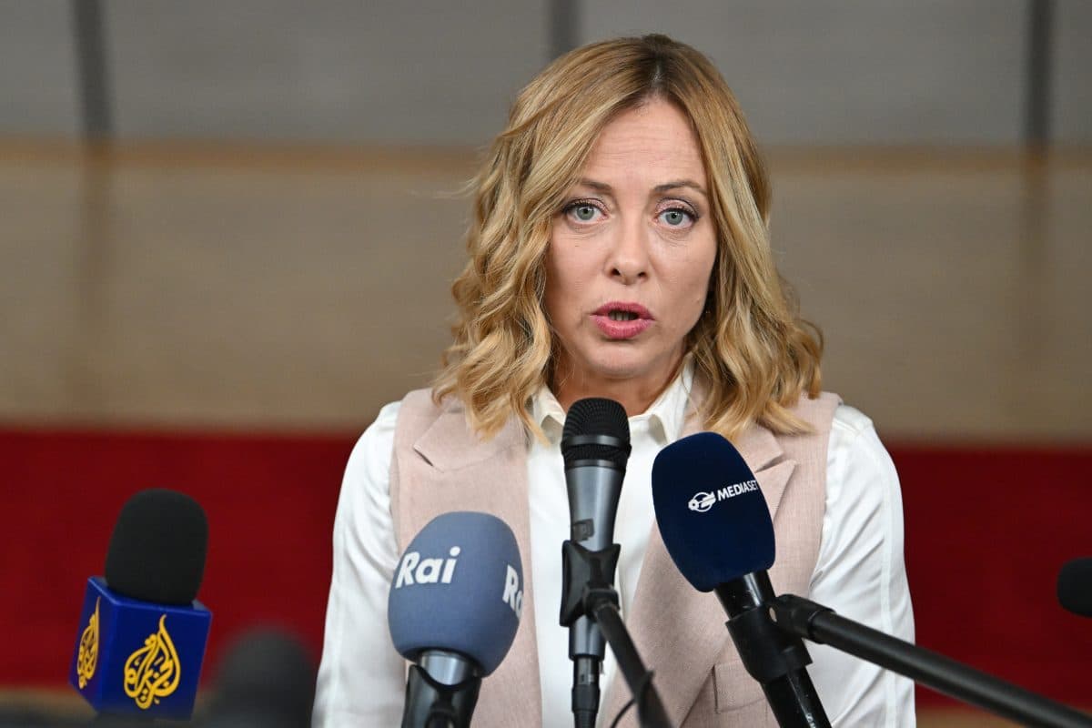 En kvinne med blondt hår snakker på en pressekonferanse, stående bak flere mikrofoner merket Rai, Al Jazeera og Mediaset.