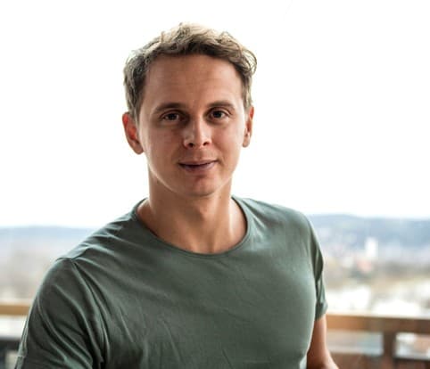 En mann med kort, lysebrunt hår og grønn t-skjorte står utendørs med en uskarp bakgrunn av trær og bygninger.