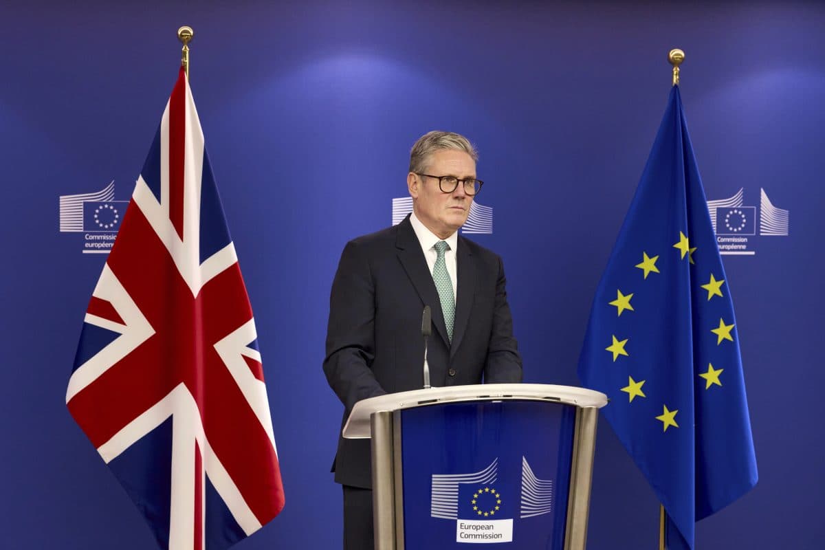 En mann i dress står på et podium mellom Storbritannias og EUs flagg, med EU-kommisjonens logoer i bakgrunnen.