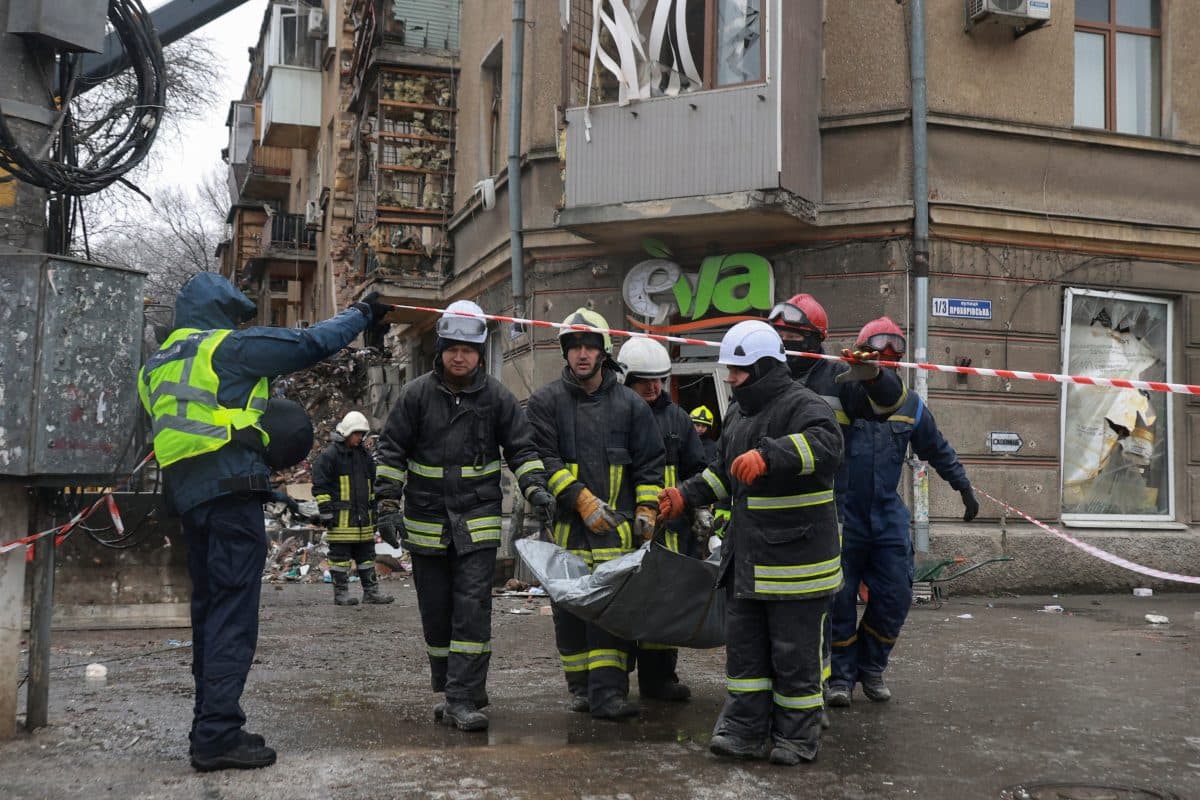 Brannmenn bærer en båre forbi en skadet bygning, med ruiner og ødeleggelser synlige rundt seg, mens en politibetjent overvåker åstedet.