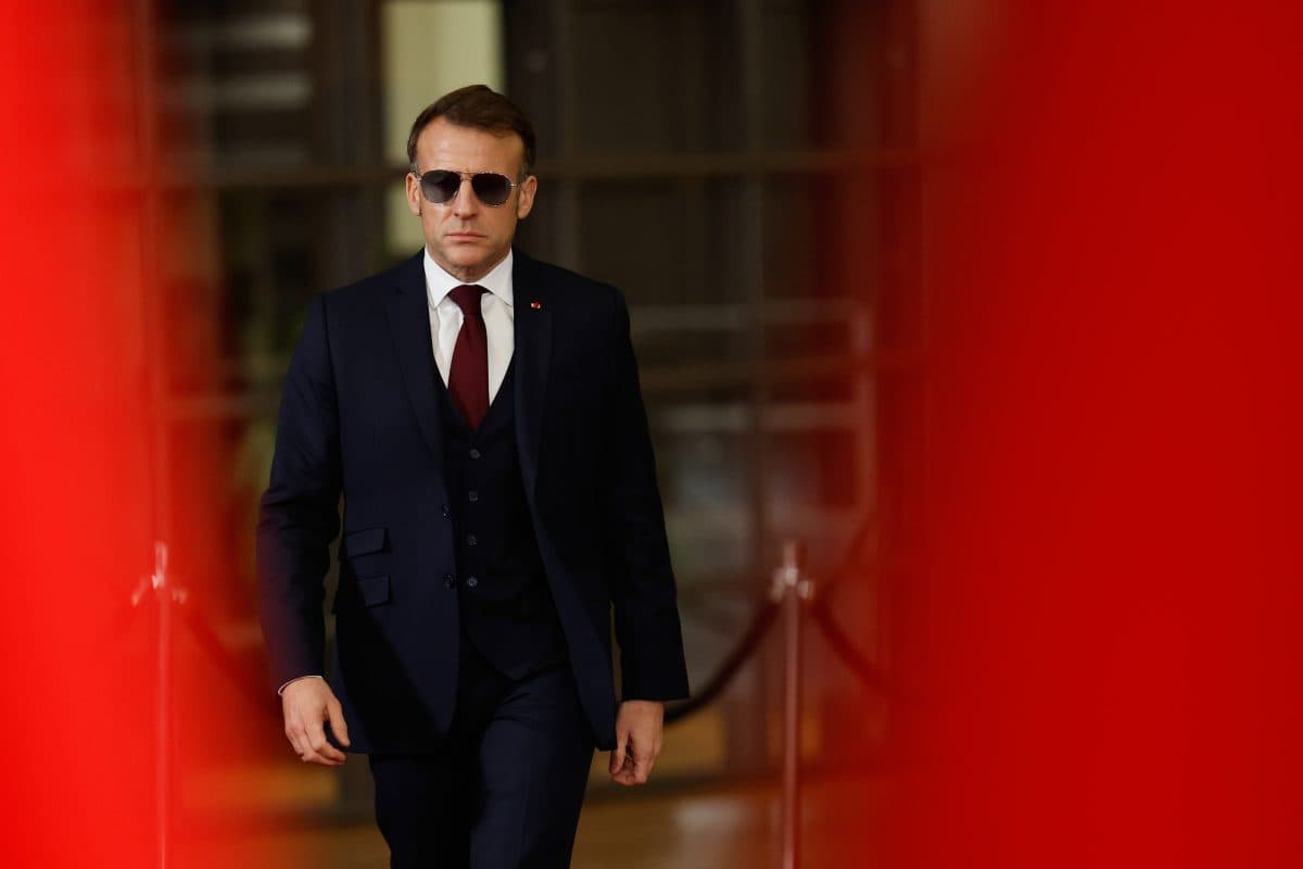 En mann i mørk dress og solbriller går fremover innendørs, innrammet av uskarpe røde former i forgrunnen.