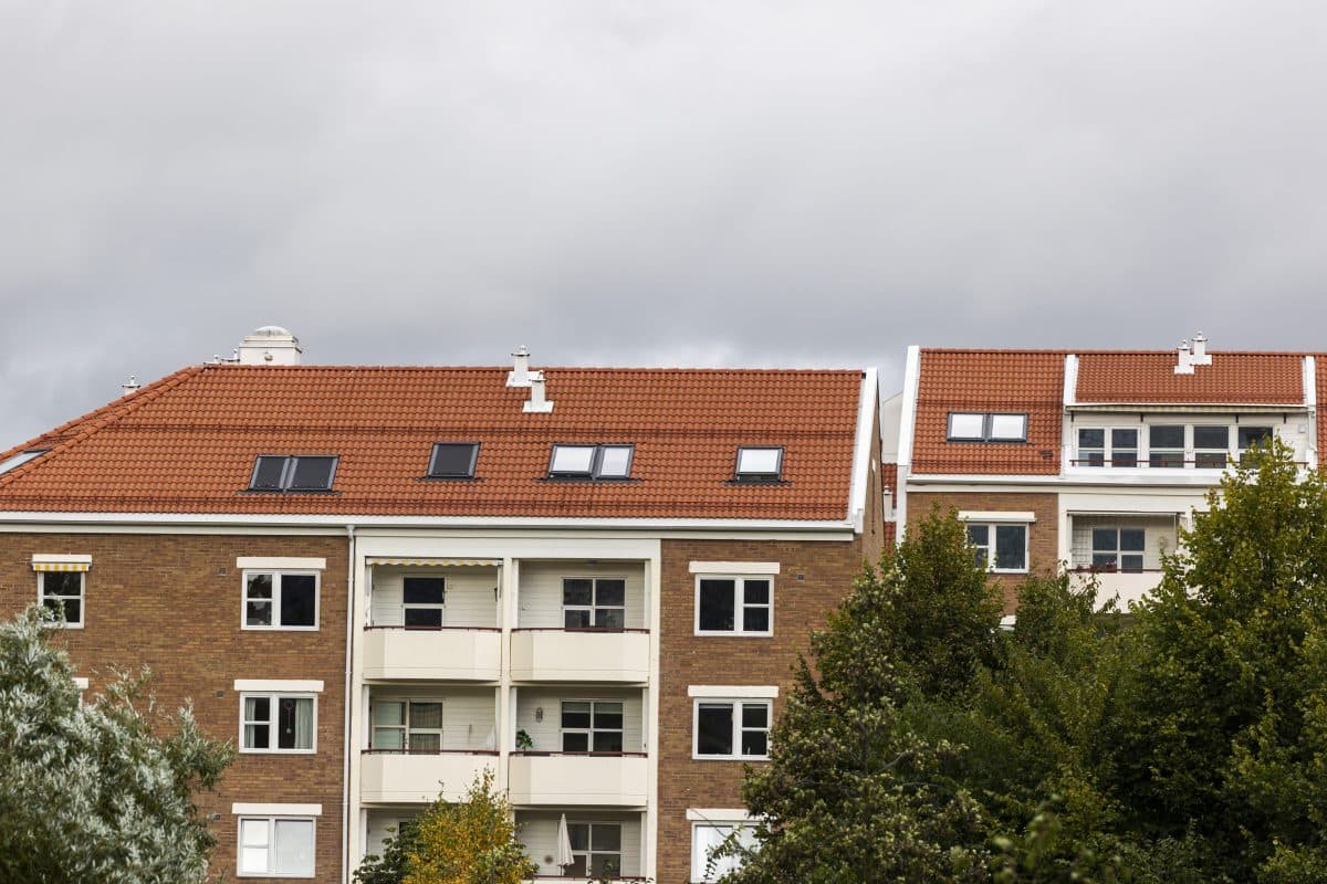 Tre boligblokker med røde tegltak og balkonger står bak grønne trær under en overskyet himmel.