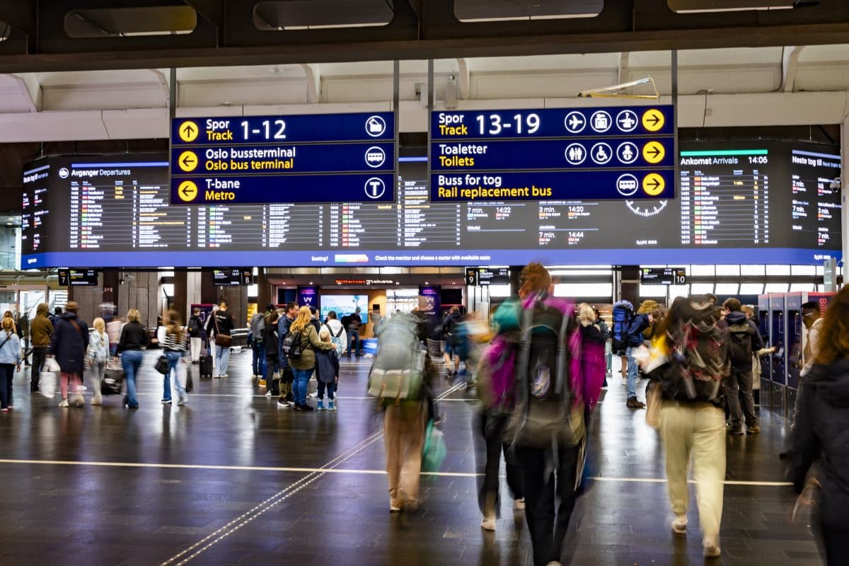 En folkemengde med ryggsekker går under store digitale avgangstavler som viser tog- og bussinformasjon på en travel stasjon.