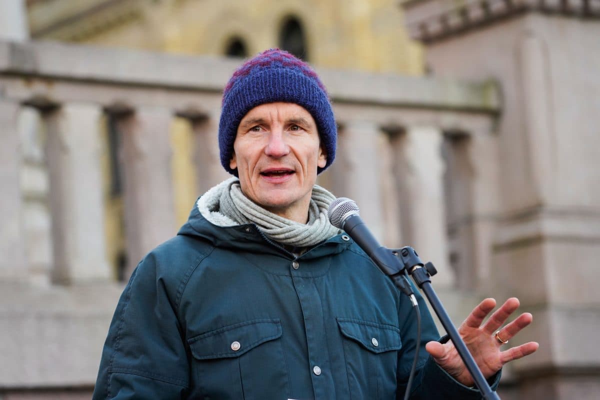 En mann i blå vinterjakke og strikkelue snakker i en mikrofon utendørs foran et steinrekkverk.