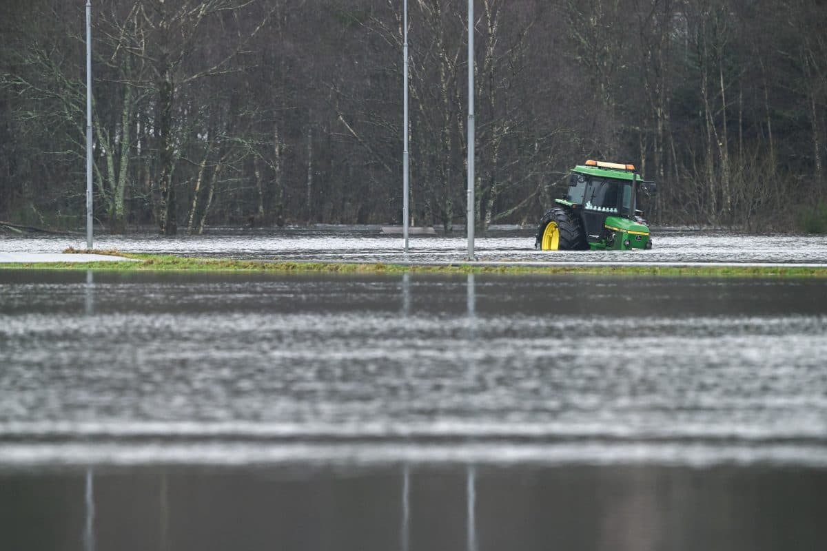 En grønn traktor står delvis under vann på et vannmettet jorde med løvløse trær i bakgrunnen.