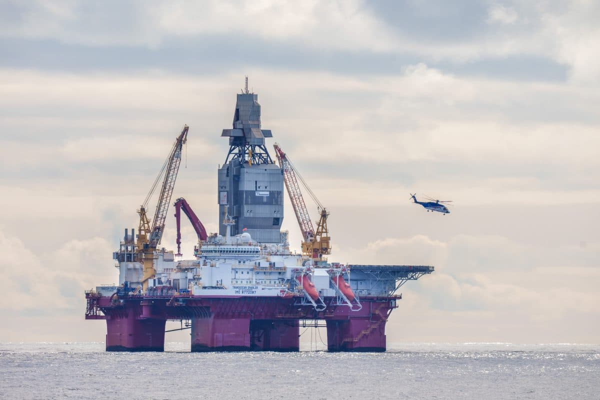 En oljeboringsplattform i havet med kraner og utstyr synlig; et helikopter flyr i nærheten under en overskyet himmel.