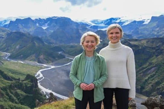 To kvinner står på en gresskledd klippe med utsikt over en bred elvedal og fjell under en delvis overskyet himmel.