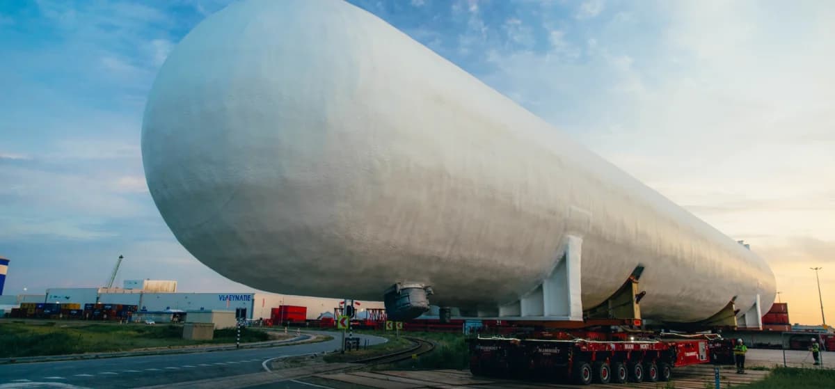 En stor, sylinderformet industritank transporteres på et kjøretøy med flere hjul langs en vei i nærheten av lagerbygninger og kraner i solnedgangen.