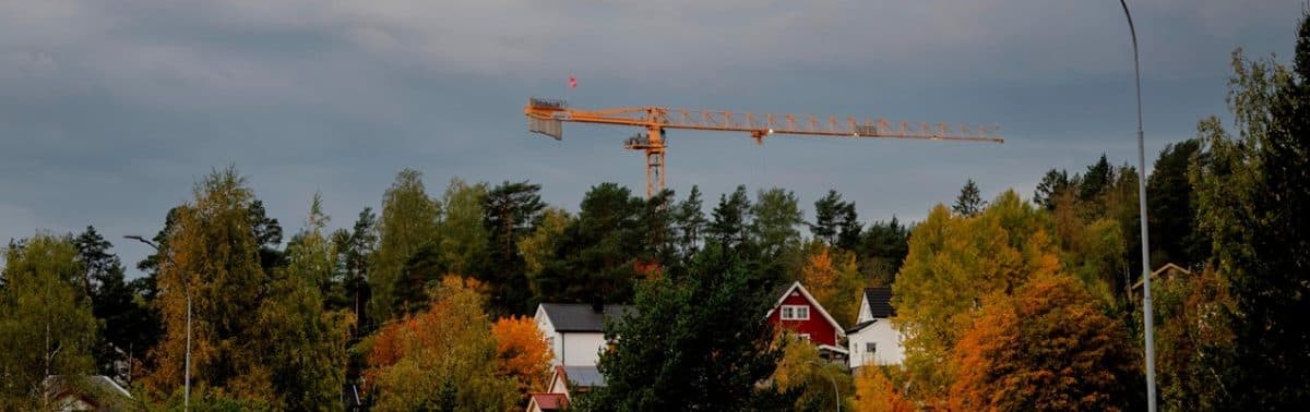En gul byggekran hever seg over hus og trær med høstløv under en overskyet himmel.
