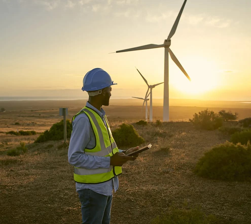 En person i sikkerhetsvest og hjelm står foran vindturbiner i solnedgangen og holder et nettbrett i et landlig landskap.