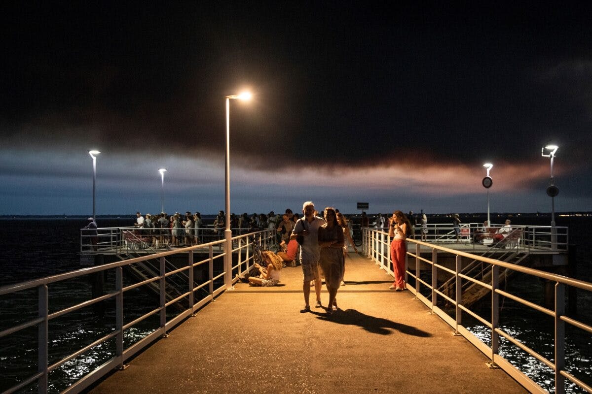 Folk går på havnepir med mørk røyksky fra skogbranner i bakgrunnen