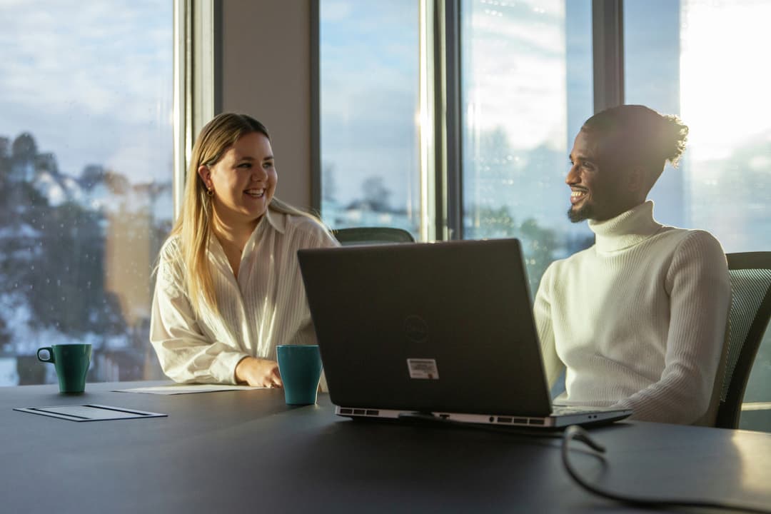 To personer sitter ved et bord med en bærbar datamaskin foran et vindu.