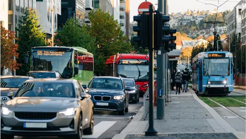 Travel bygate med biler, busser og trikk, med moderne bygninger og hus i åssiden i bakgrunnen.