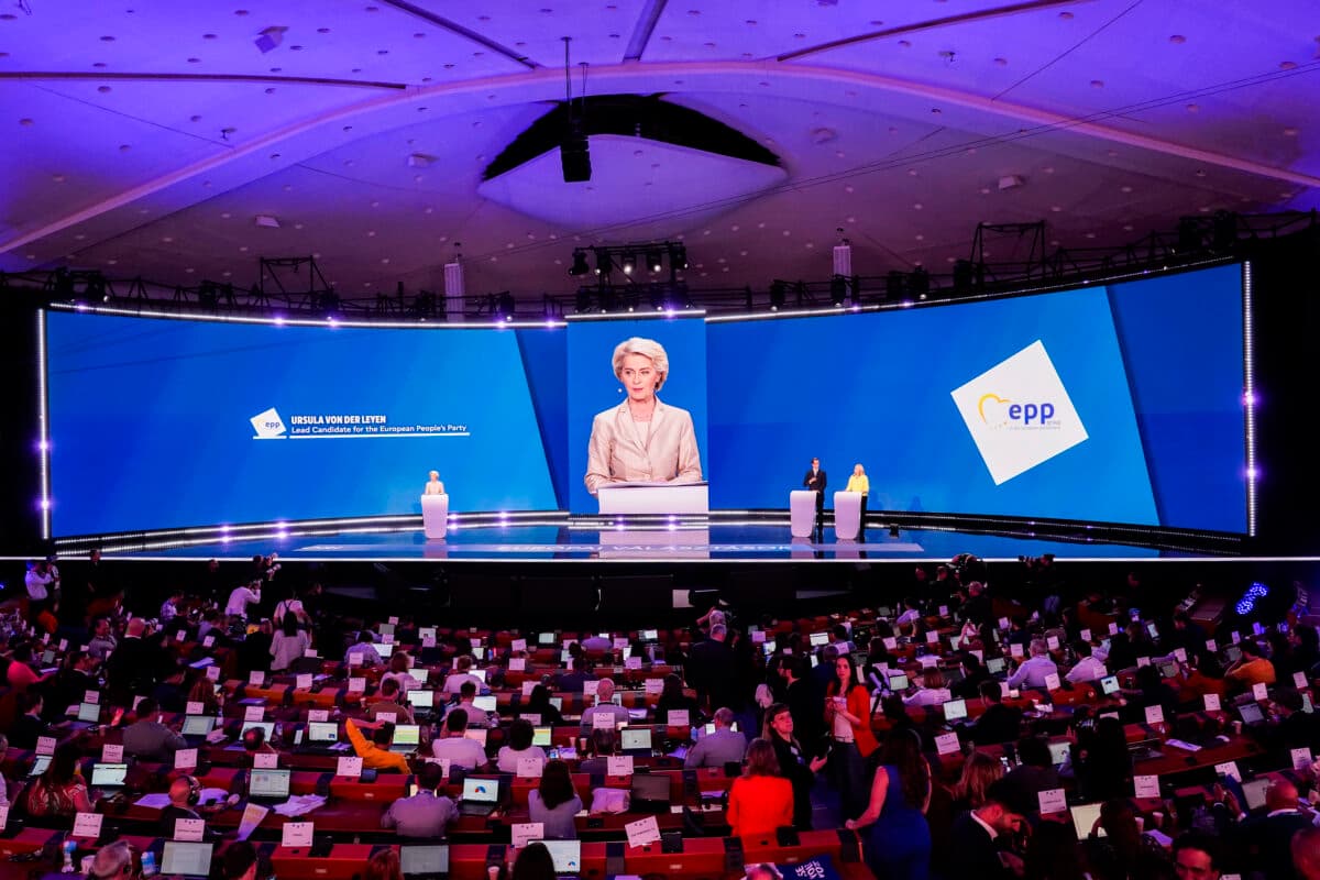 En foredragsholder står på et podium på en stor scene med blå skjermer som viser logoer, og henvender seg til et publikum som sitter med bærbare datamaskiner i en romslig konferansesal.