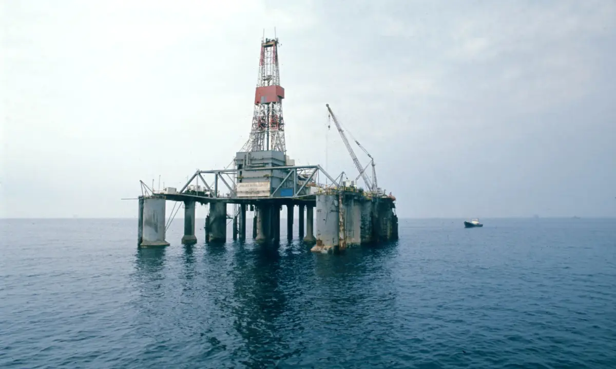 En borerigg står midt i et stille hav under en overskyet himmel, med en liten båt synlig i det fjerne.