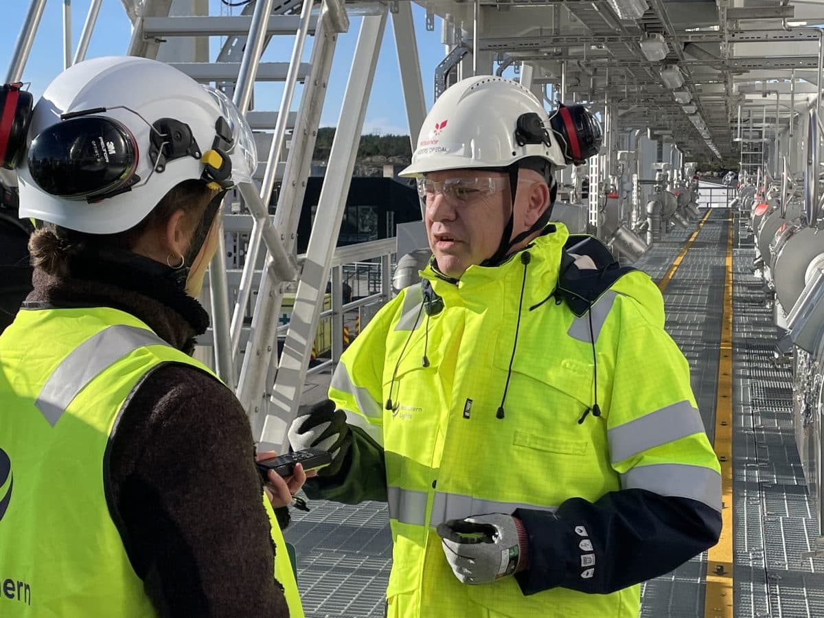 To bygningsarbeidere i synlige jakker og hjelmer snakker på en metallgangvei. Industrielt utstyr er i bakgrunnen.