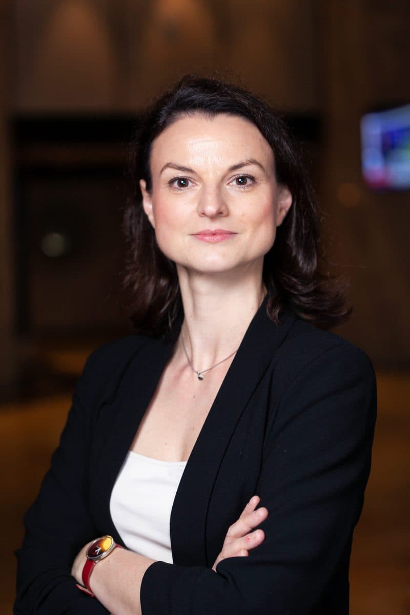 En person med skulderlangt brunt hår, iført svart blazer og hvit topp, står med armene i kors i et svakt opplyst innendørsmiljø.