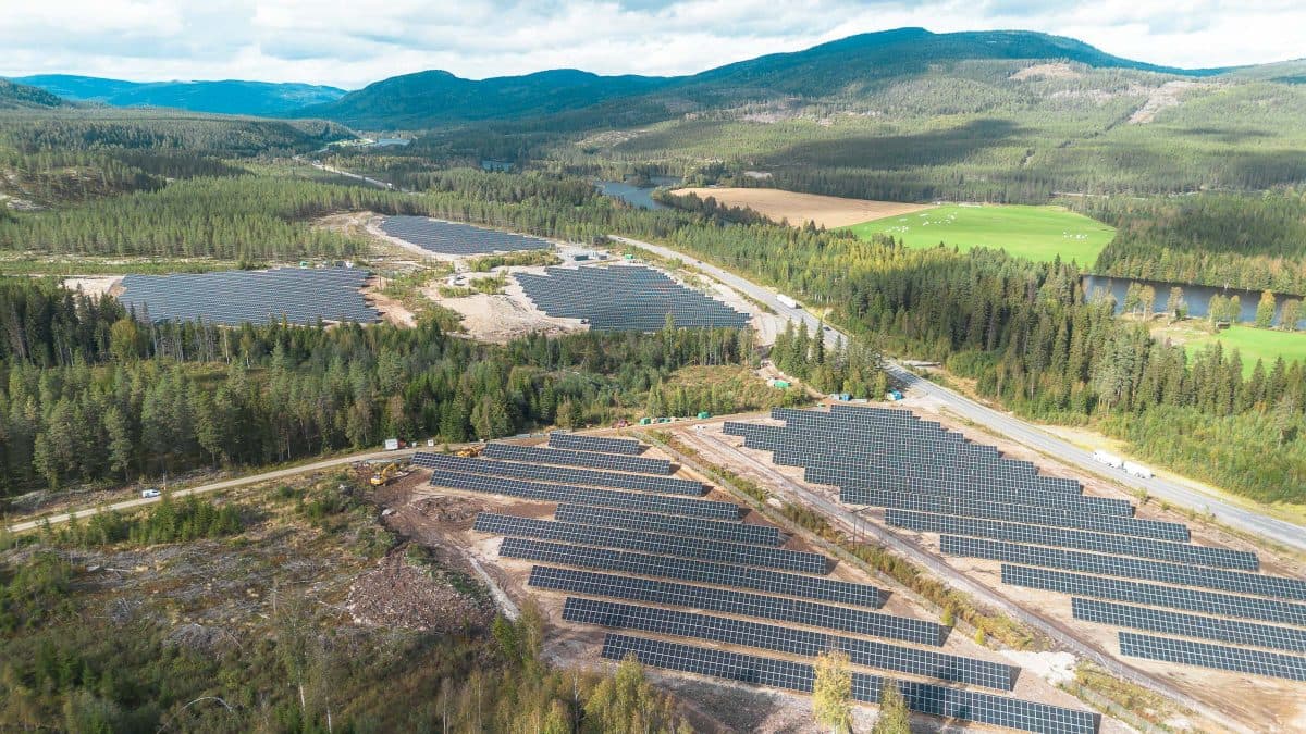 Luftfoto av ekspansive solcellepaneler midt i et frodig, grønt landskap med åser og trær under en delvis overskyet himmel.