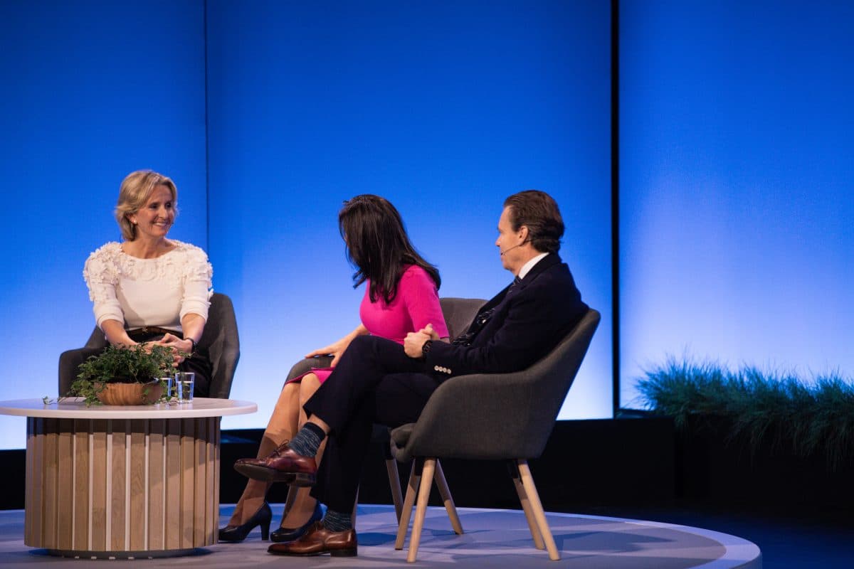 Tre personer som sitter på scenen har en samtale; to kvinner og en mann i forretningsantrekk, med blått bakteppe og et lite bord med planter foran seg.