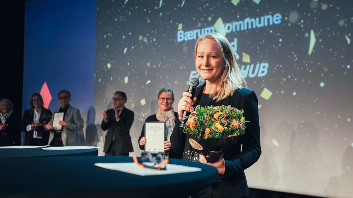 En kvinne som holder en bukett står på scenen, smiler og snakker i en mikrofon. Flere personer i bakgrunnen applauderer mens de holder plaketter. Et dekorativt banner vises bak dem.