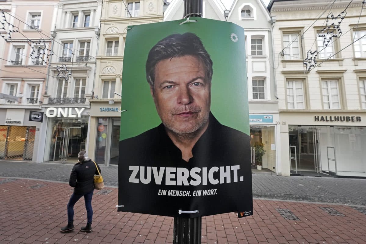En politisk plakat på en gate viser en mann mot grønn bakgrunn med teksten "ZUVERSICHT. EIN MENSCH. EIN WORT." Fotgjengere og butikker er i bakgrunnen.