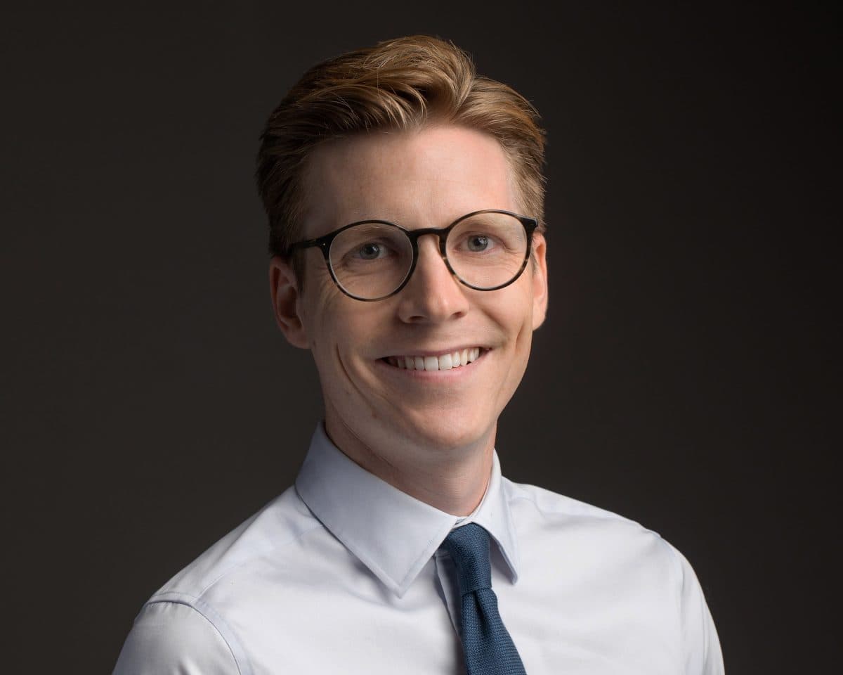 Person med kort hår, iført briller, hvit skjorte og blått slips, smilende foran en mørk bakgrunn.