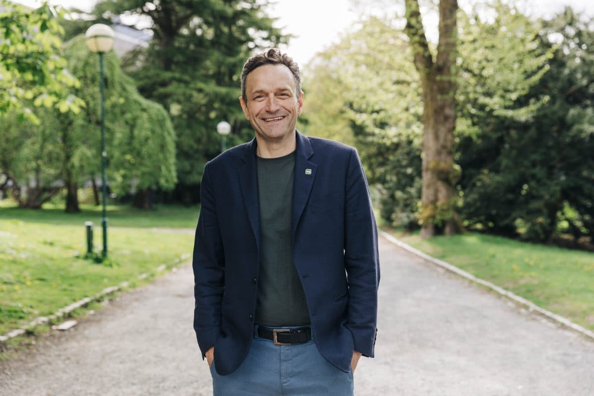 En person som står på en grussti i en park, iført marineblazer og grønn skjorte, med trær og grønt i bakgrunnen.