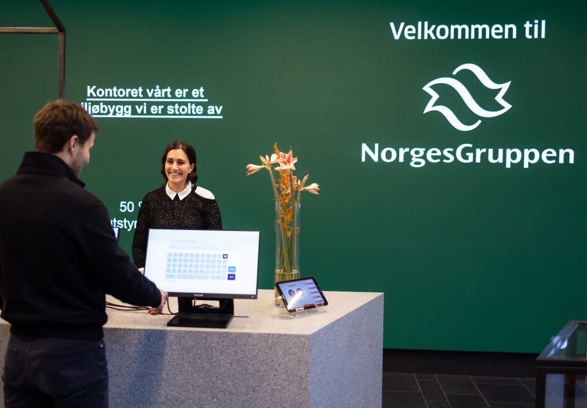 En mann står ved en resepsjon og kommuniserer med en smilende kvinnelig resepsjonist. Bakteppet har en grønn vegg med "Velkommen til NorgesGruppen" og blomsterdekorasjoner.