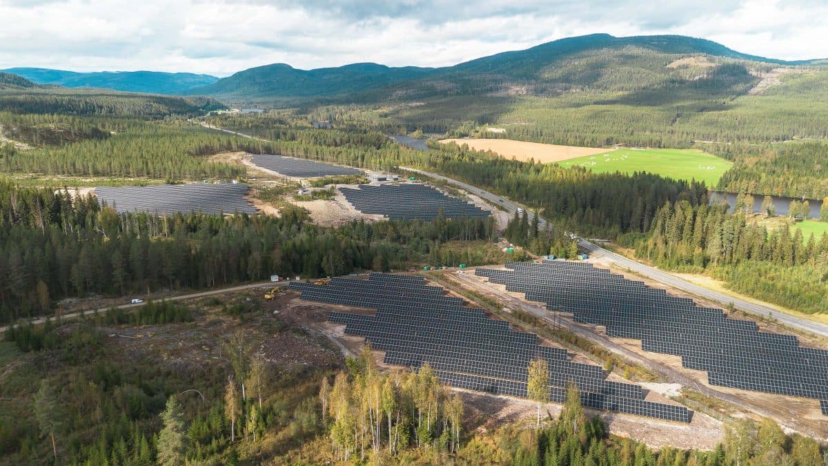Luftfoto av flere solcellepanelinstallasjoner i et skogkledd, fjellrikt landskap under en delvis overskyet himmel.
