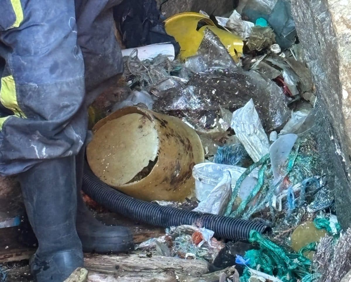 En person i svarte støvler står ved siden av en haug med diverse søppel, inkludert ødelagte bøtter, plast og rusk.