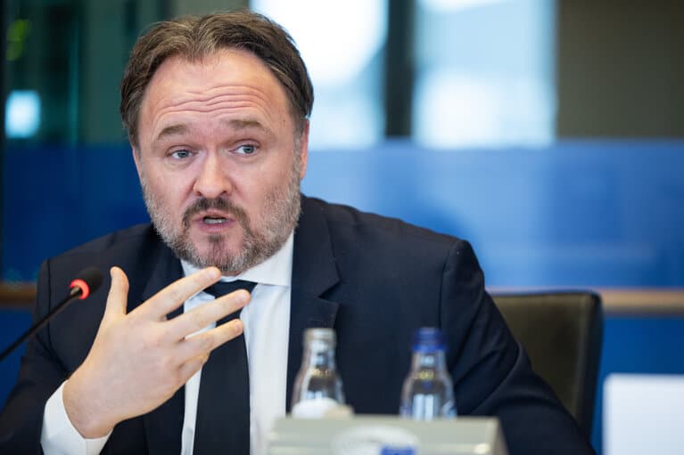 En mann i dress og slips snakker inn i en mikrofon ved et konferansebord og gestikulerer med hånden. Vannflasker er synlige på bordet foran ham.