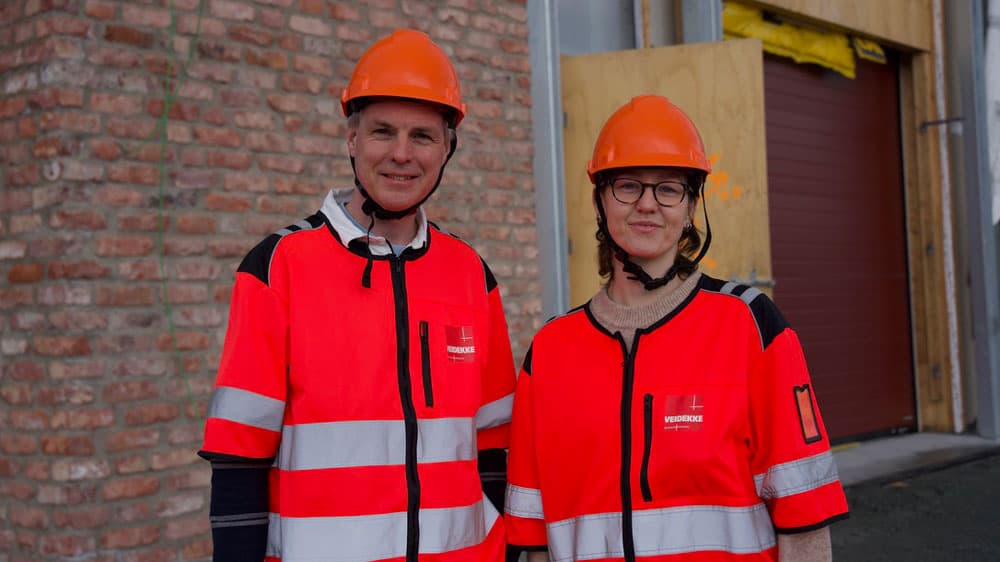 To personer med oransje sikkerhetshjelmer og refleksvester med høy synlighet står foran en murvegg og en bygning under bygging.