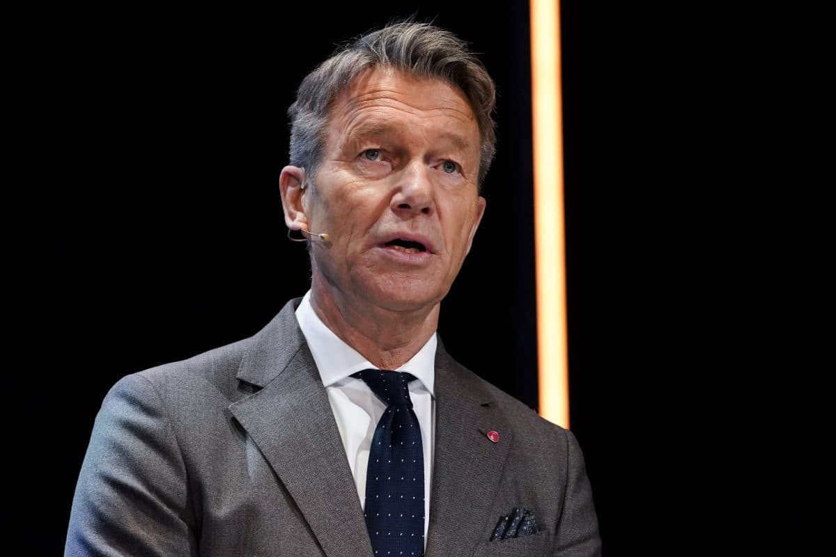 En mann i grå dress og marineblått slips snakker mens han har på seg en headsetmikrofon, stående mot en mørk bakgrunn med et vertikalt oransje lys.