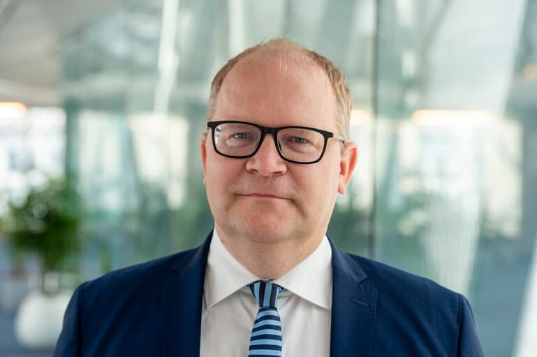 En middelaldrende mann med briller og vikende hårfeste, iført dress og stripete slips, står i et innemiljø med uskarpt glass og planter i bakgrunnen.