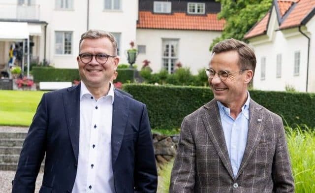 To menn i dress går på en grussti utenfor et stort, hvitt hus omgitt av grønne planter, med hver sin papirrull i hånden og et smil om munnen.