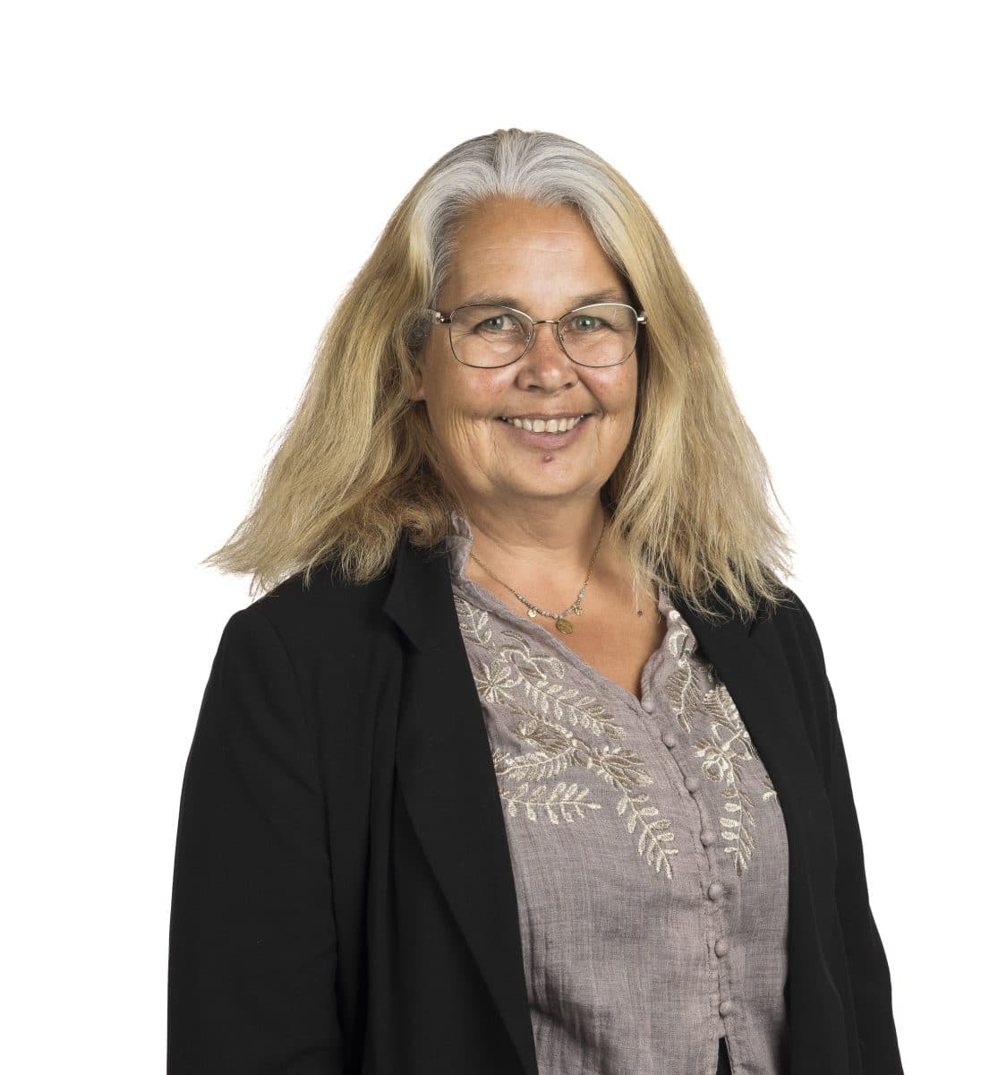 En kvinne med langt blondt hår, iført briller, svart blazer og en lys, brodert bluse, smiler foran en hvit bakgrunn.