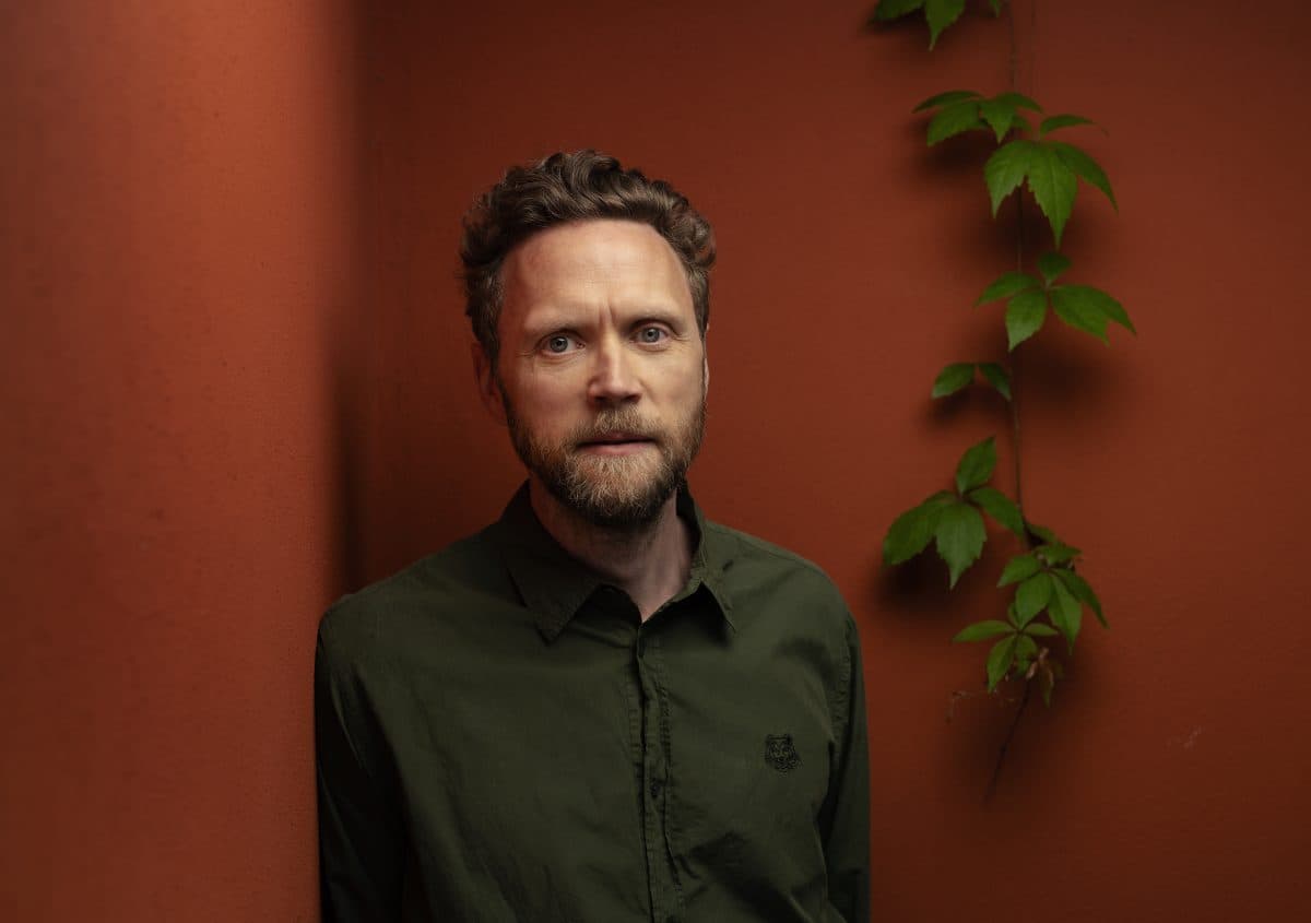 En mann med kort, krøllete hår og skjegg står mot en rustfarget vegg med en grønn vinranke som klatrer ved siden av ham. Han har på seg en grønn skjorte med knapper.