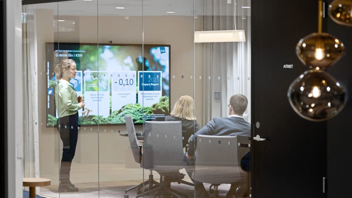 En person står og presenterer data på en skjerm for to sittende kolleger i et moderne konferanserom med glassvegger.