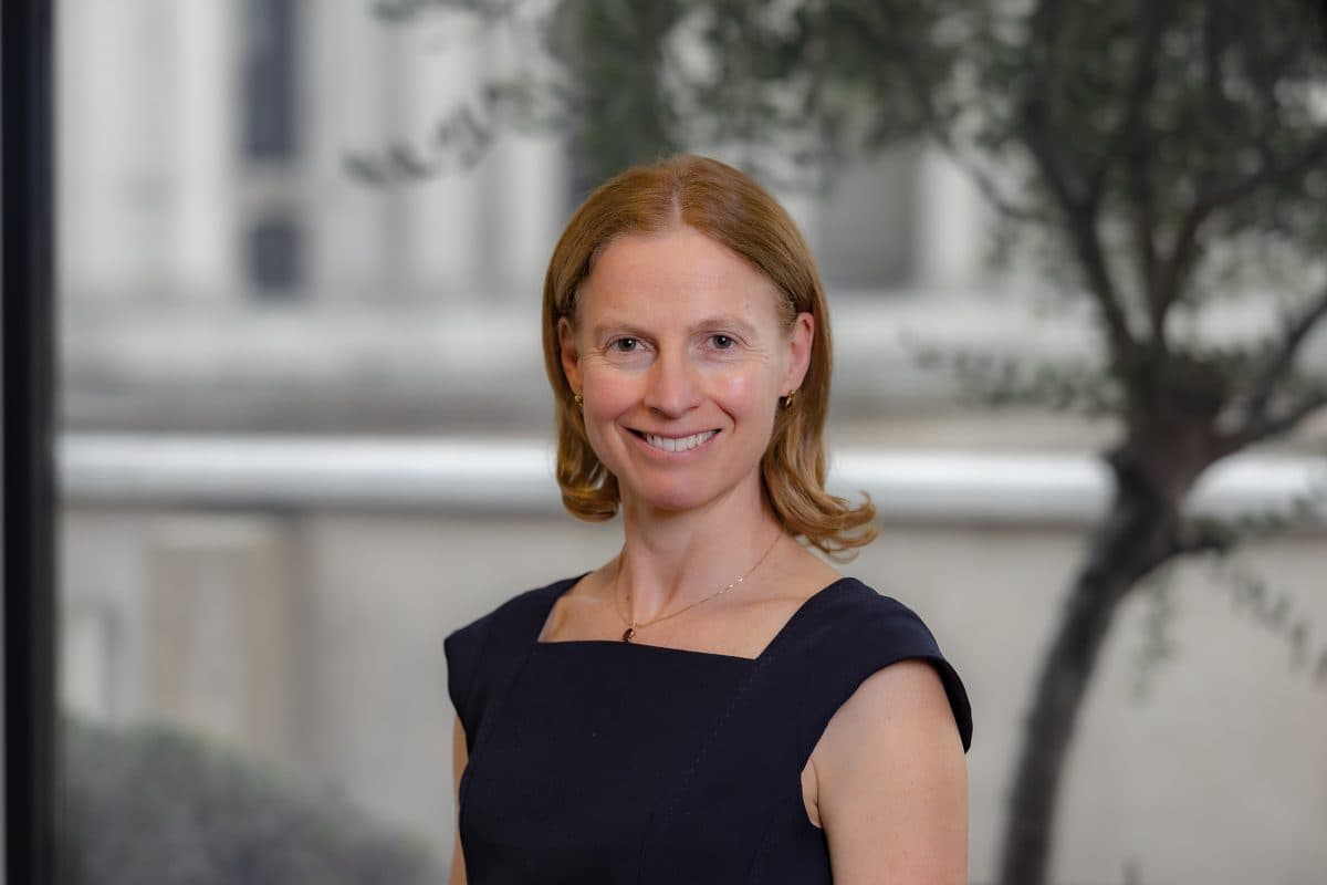 En kvinne med skulderlangt, rødlig hår, iført en mørk, ermeløs kjole, står smilende foran en uskarp utendørs bakgrunn med trær.