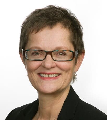 En kvinne med kort, brunt hår, briller og svart blazer smiler til kameraet mot en enkel, hvit bakgrunn.